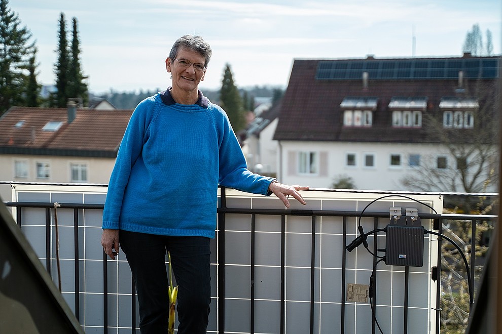 Eine ältere Dame steht im blauen Pullover auf ihrem Balkon mit einer PV-Anlage.